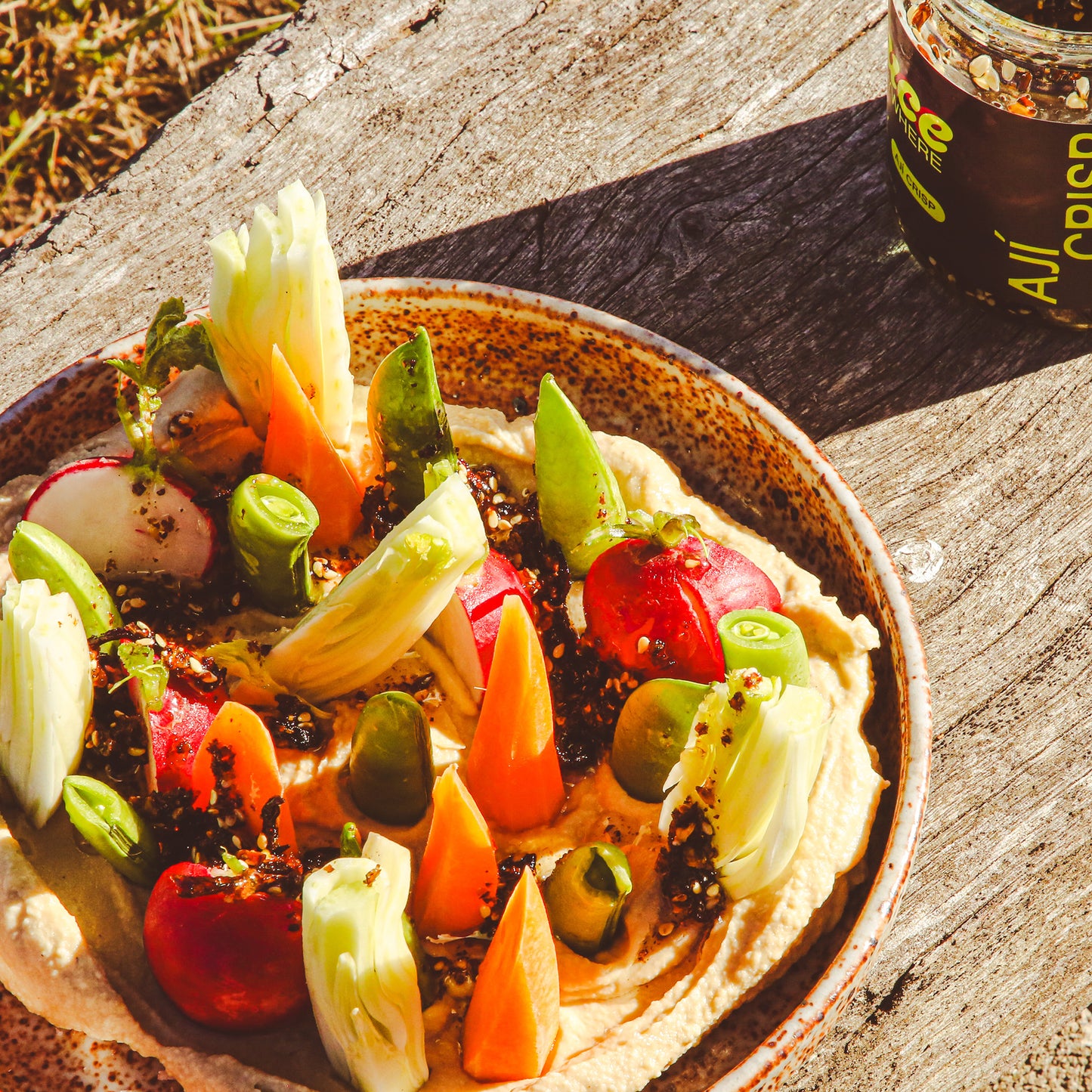 Hummus and vegetables with chile sauce in dish beside sauce jar on outdoor bench.