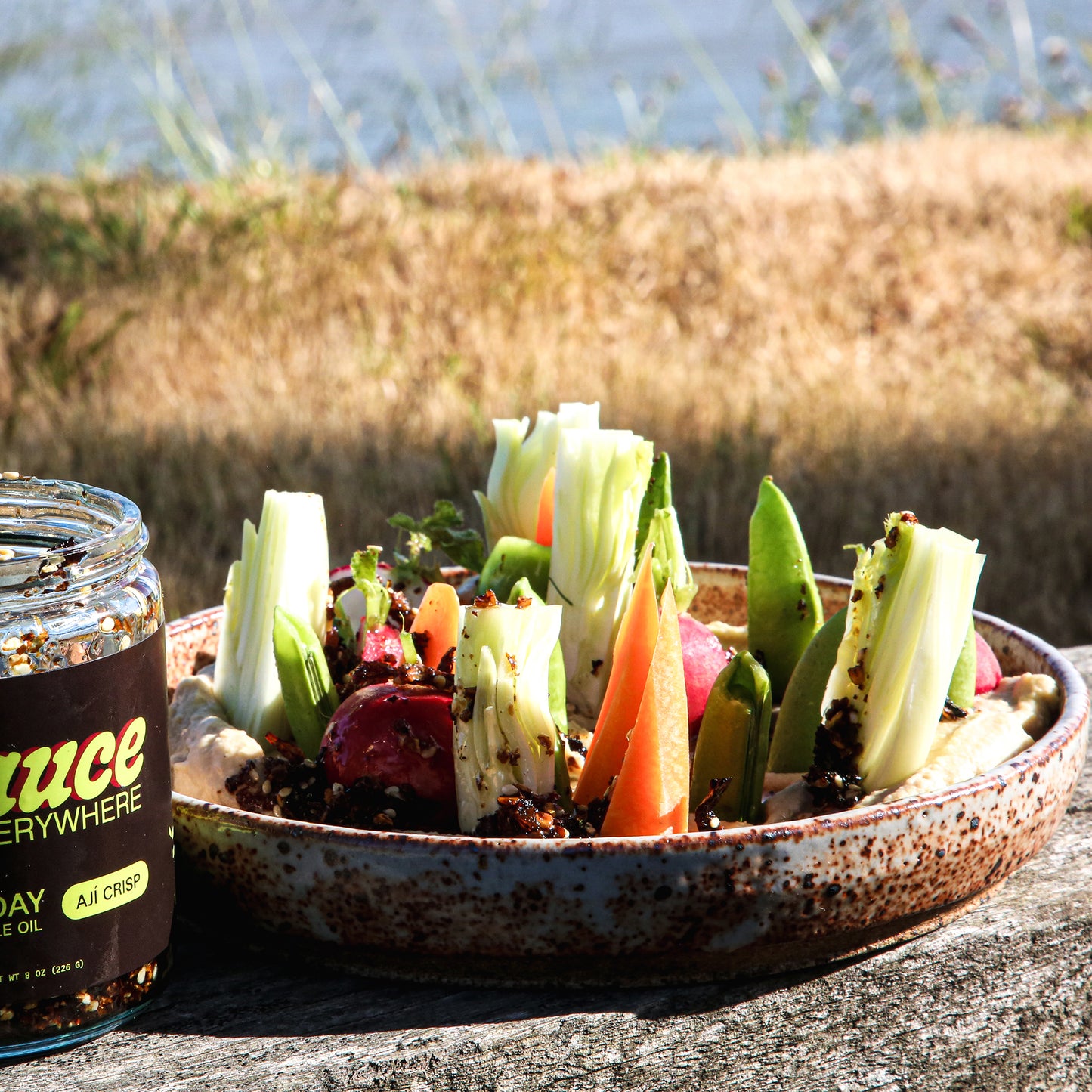 Close-up of hummus dish with vegetables and chile sauce beside sauce jar in grassy area.