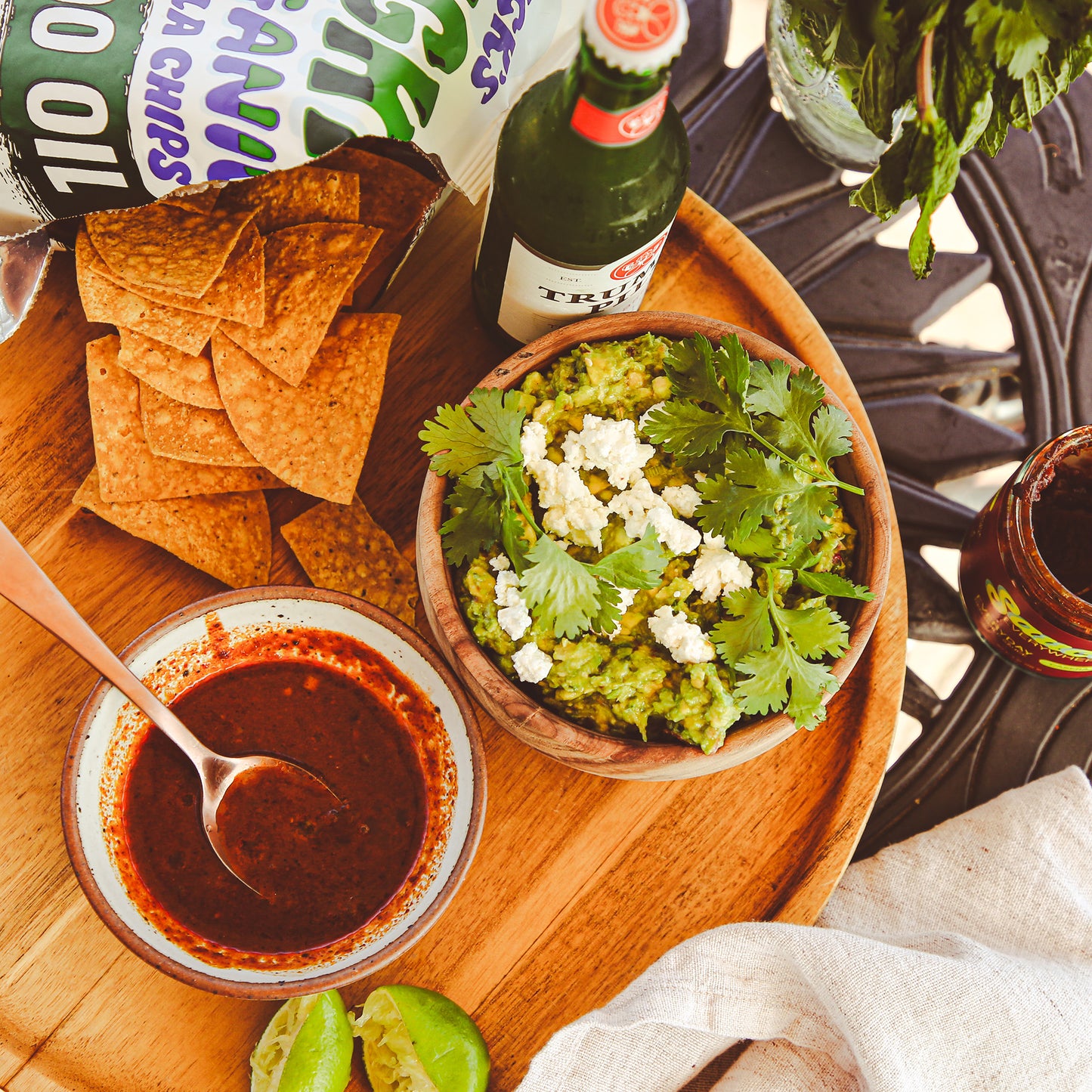 Wooden serving tray with chips, guacamole, and chile sauce on patio table.