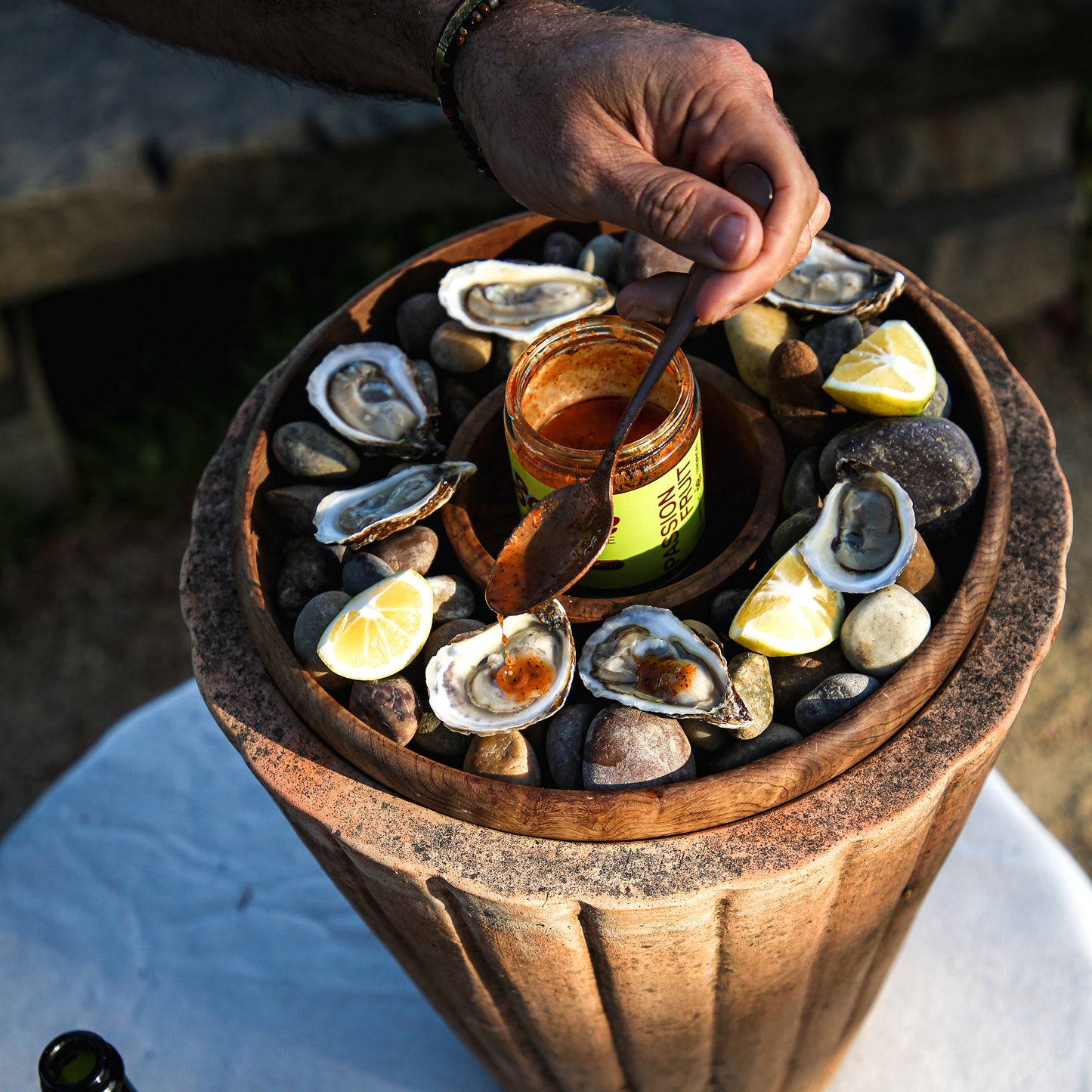Hand pouring chile sauce from spoon onto oysters in wooden tray outside.