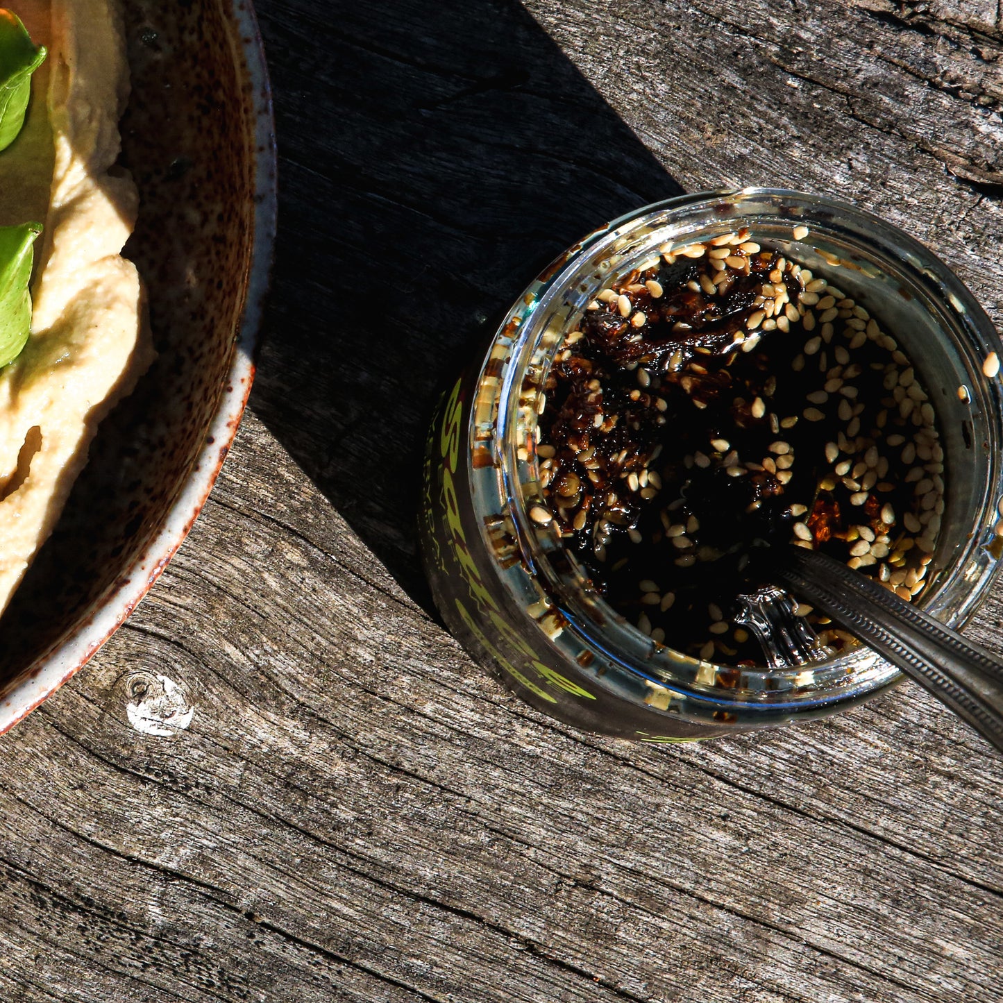 Chile sauce with sesame seeds in open sauce jar with spoon on wooden surface.