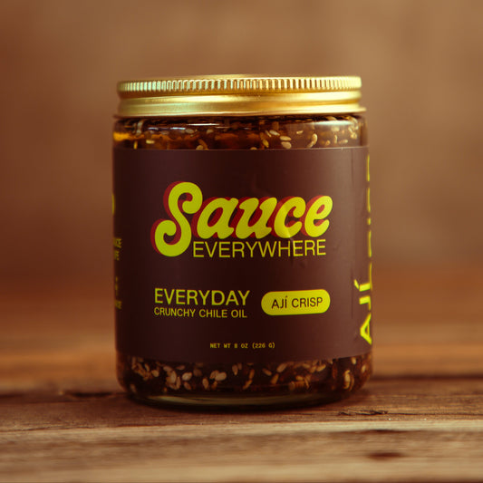 Glass sauce jar of Aji Crisp chile sauce by Sauce Everywhere on wooden table.