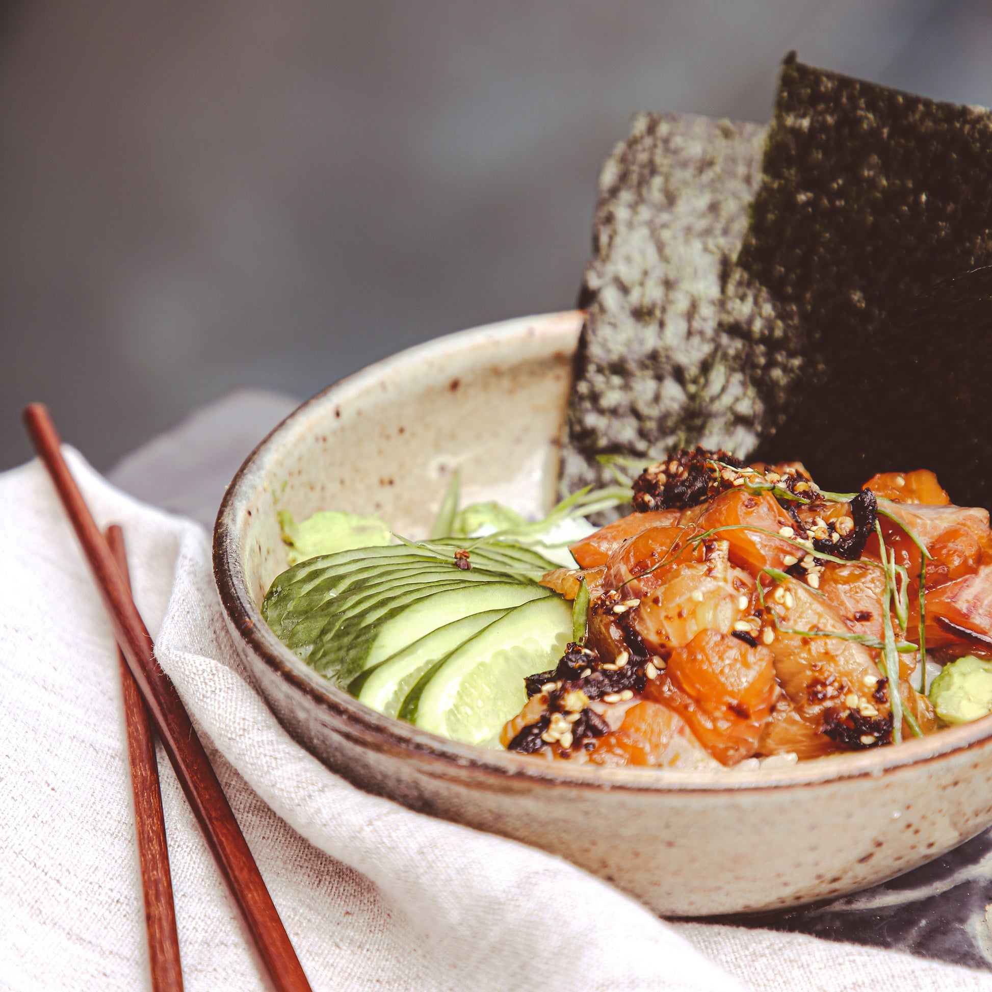 Raw trout and cucumber with chile sauce in bowl beside chopsticks and napkin.