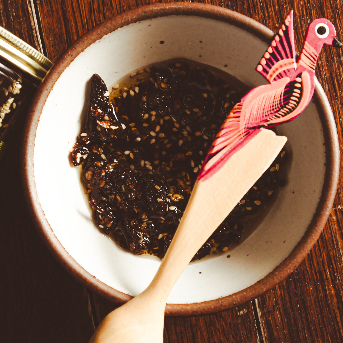Aji Crisp sauce in side bowl beside glass jar and ornamental wooden spoon.