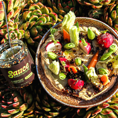 Hummus and vegetables dish beside chile sauce jar on top of vibrant succulents.