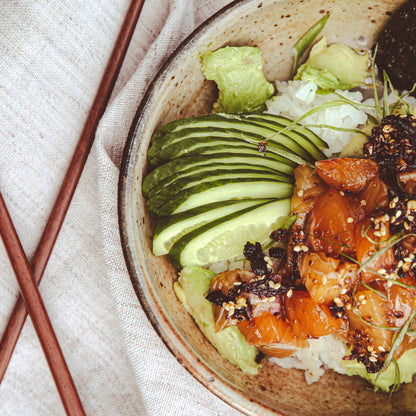 Close-up of poke bowl with chile sauce beside chopsticks and taupe napkin.