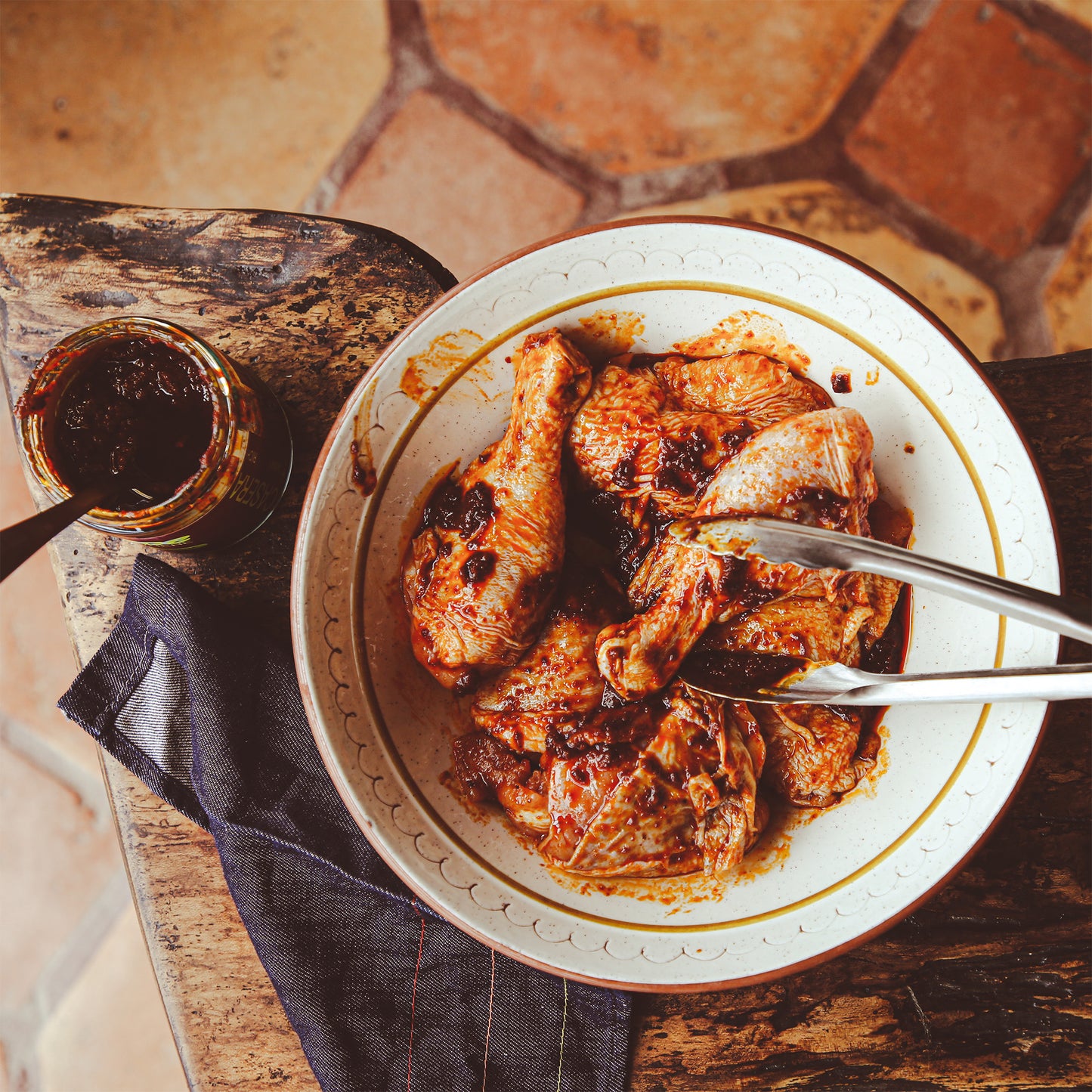 Chicken marinated with chile sauce in large bowl with tongs on wooden table.