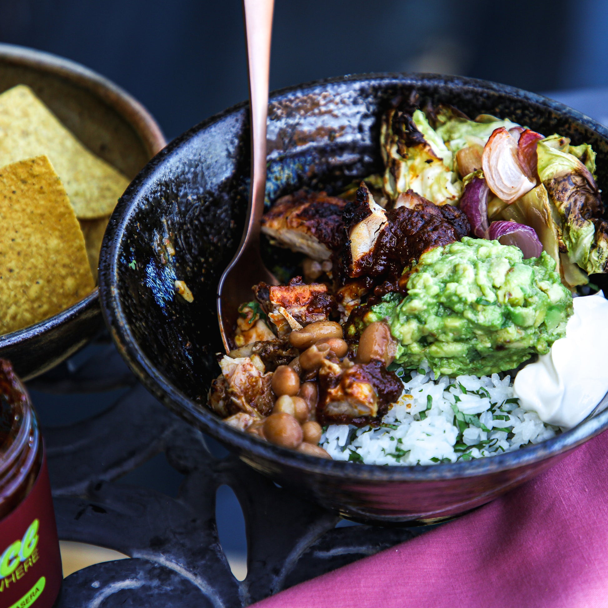 Roasted chicken, rice, beans, toppings, and chile sauce in bowl with spoon.