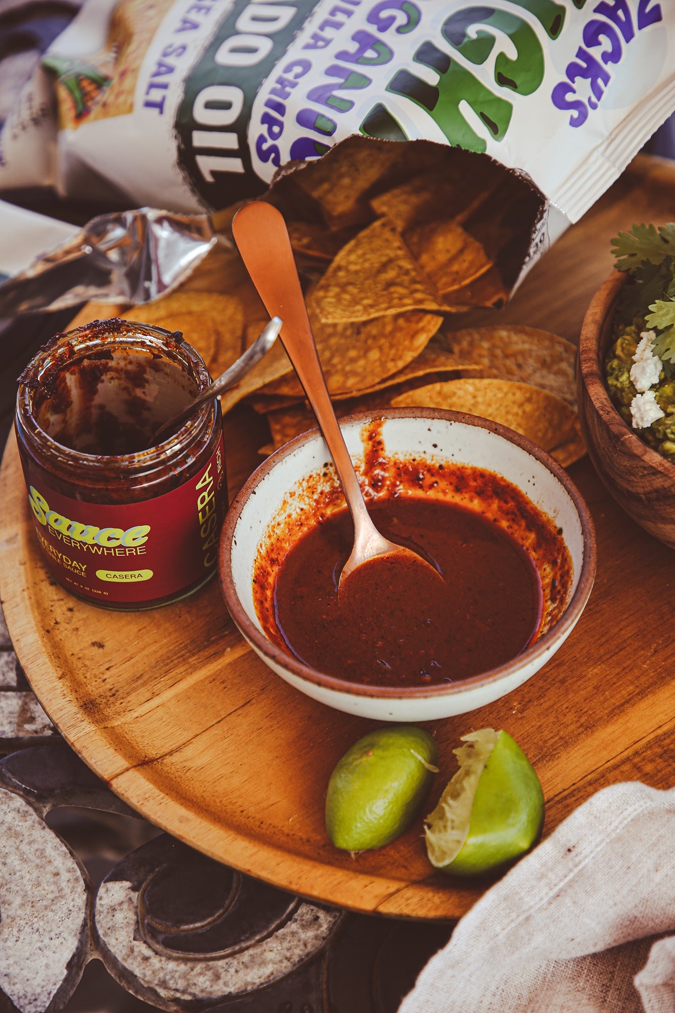 Bowl of chile salsa on wooden serving tray with chips, guacamole, and open sauce jar.