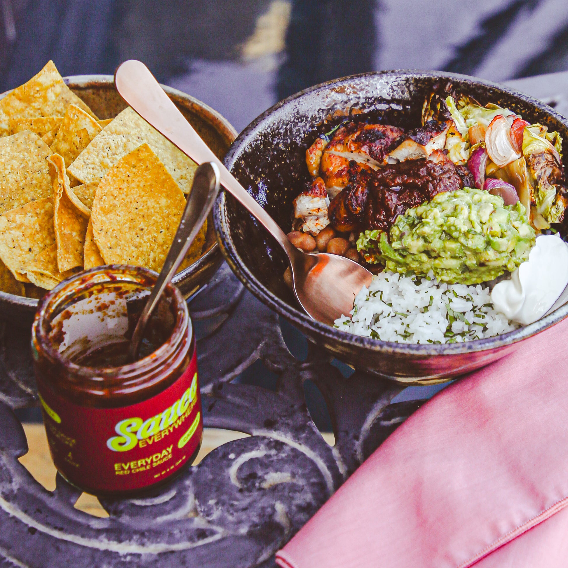 Taco bowl with chile sauce beside tortilla chips and sauce jar on metal table by pool.