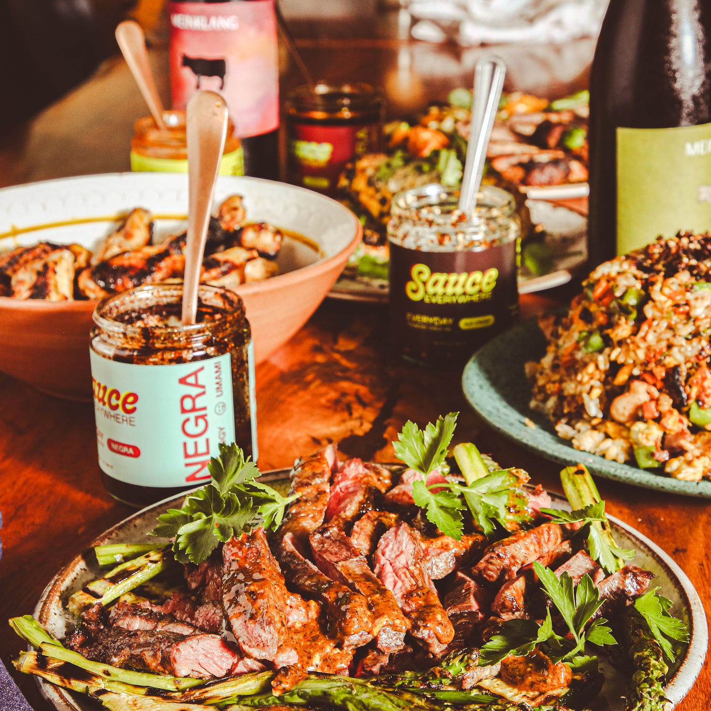 Plates of cooked meats among glass sauce jars and wine bottles on wooden table. 
