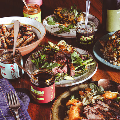 Dishes of cooked meats and vegetables among jars of chile sauce on wooden table.