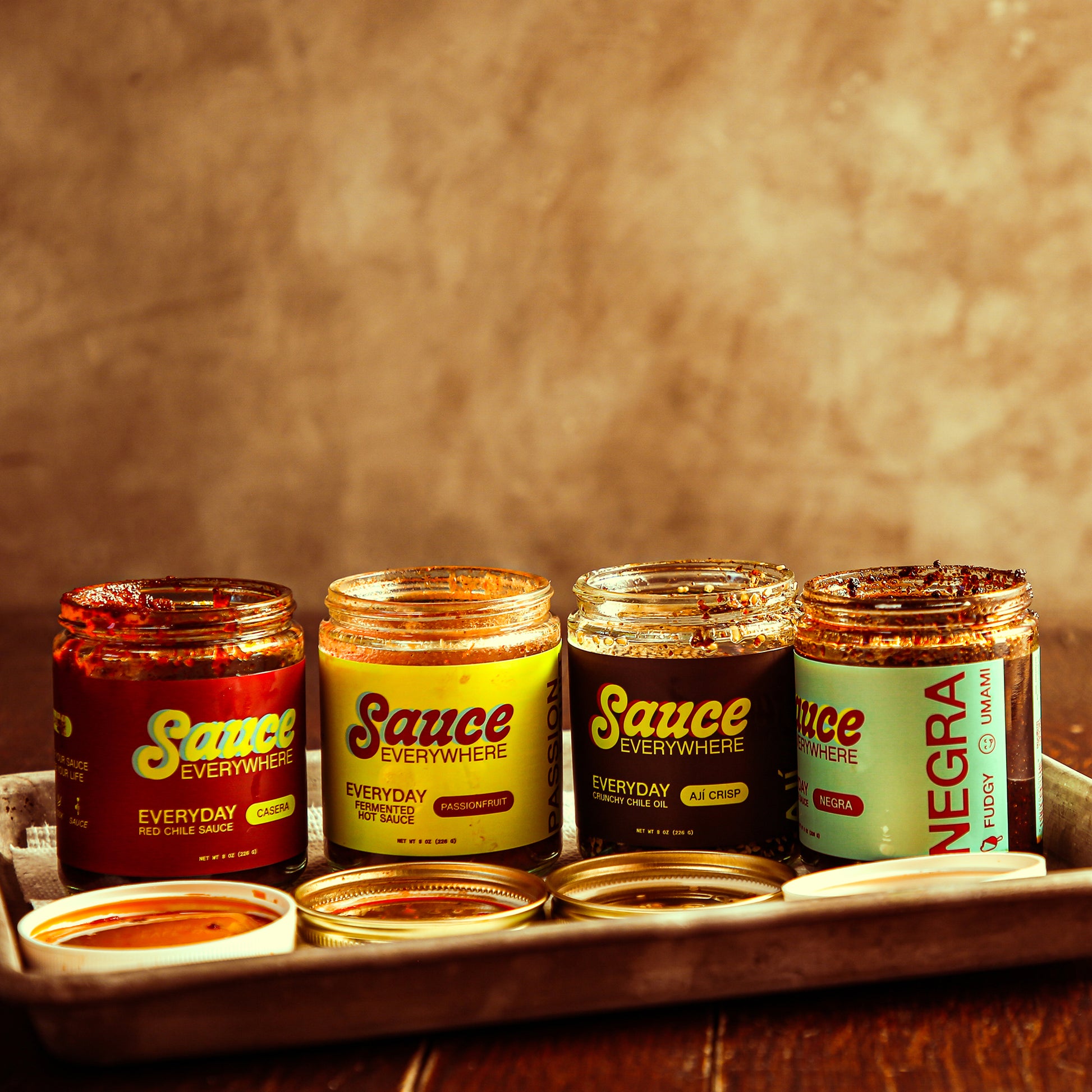 Four opened glass jars of chile sauce on metal tray on wooden table.