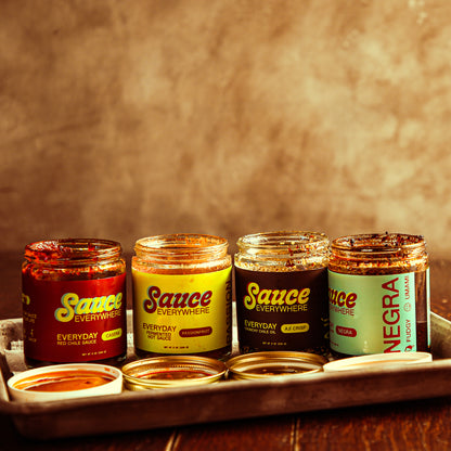 Four opened glass jars of chile sauce on metal tray on wooden table.