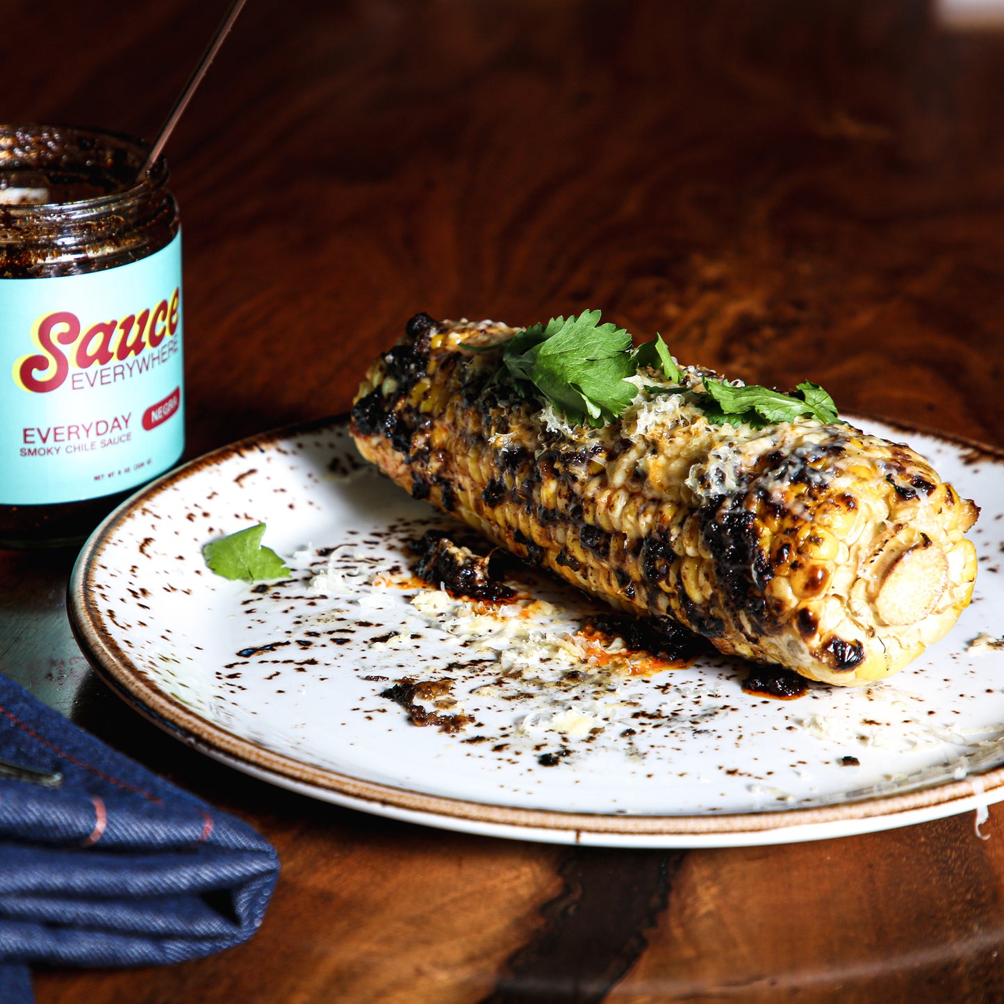 Grilled corncob with toppings and chile sauce on salad plate beside sauce jar.