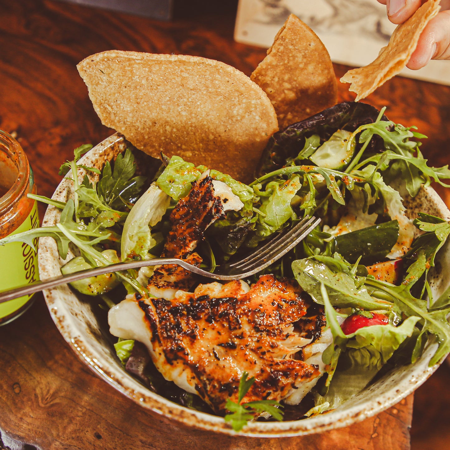 Hand lifting tostada wedge from bowl of salad with trout and chile sauce.