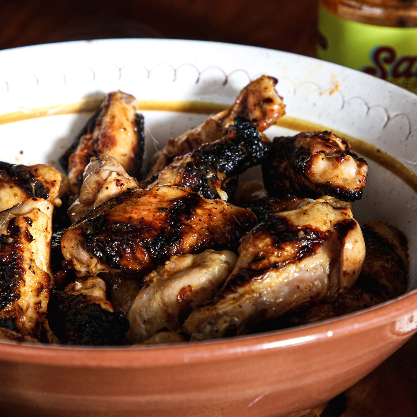 Grilled chicken wings in large bowl on table in front of sauce jar.