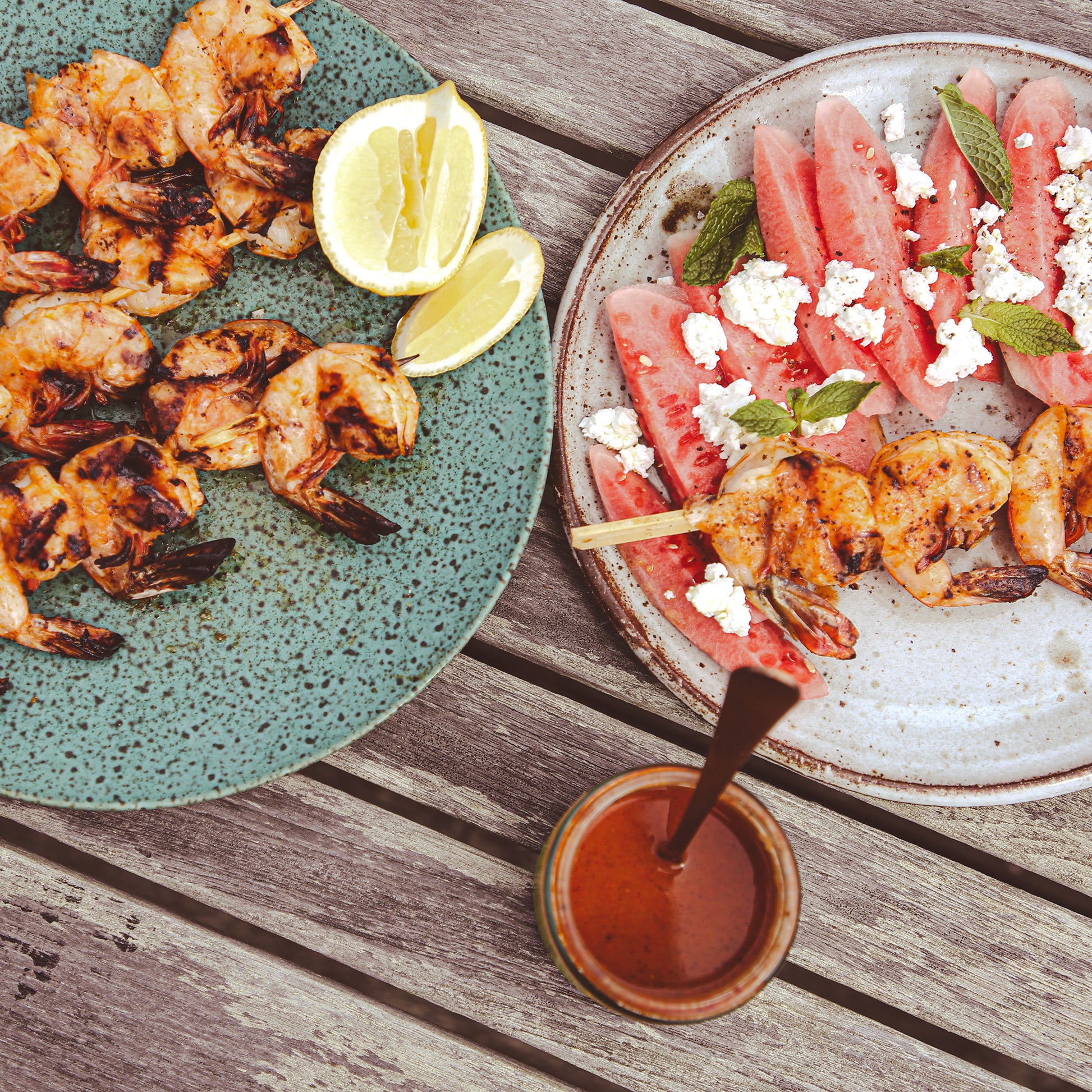 Grilled shrimp on skewers, watermelon slices, and sauce jar on outdoor table.