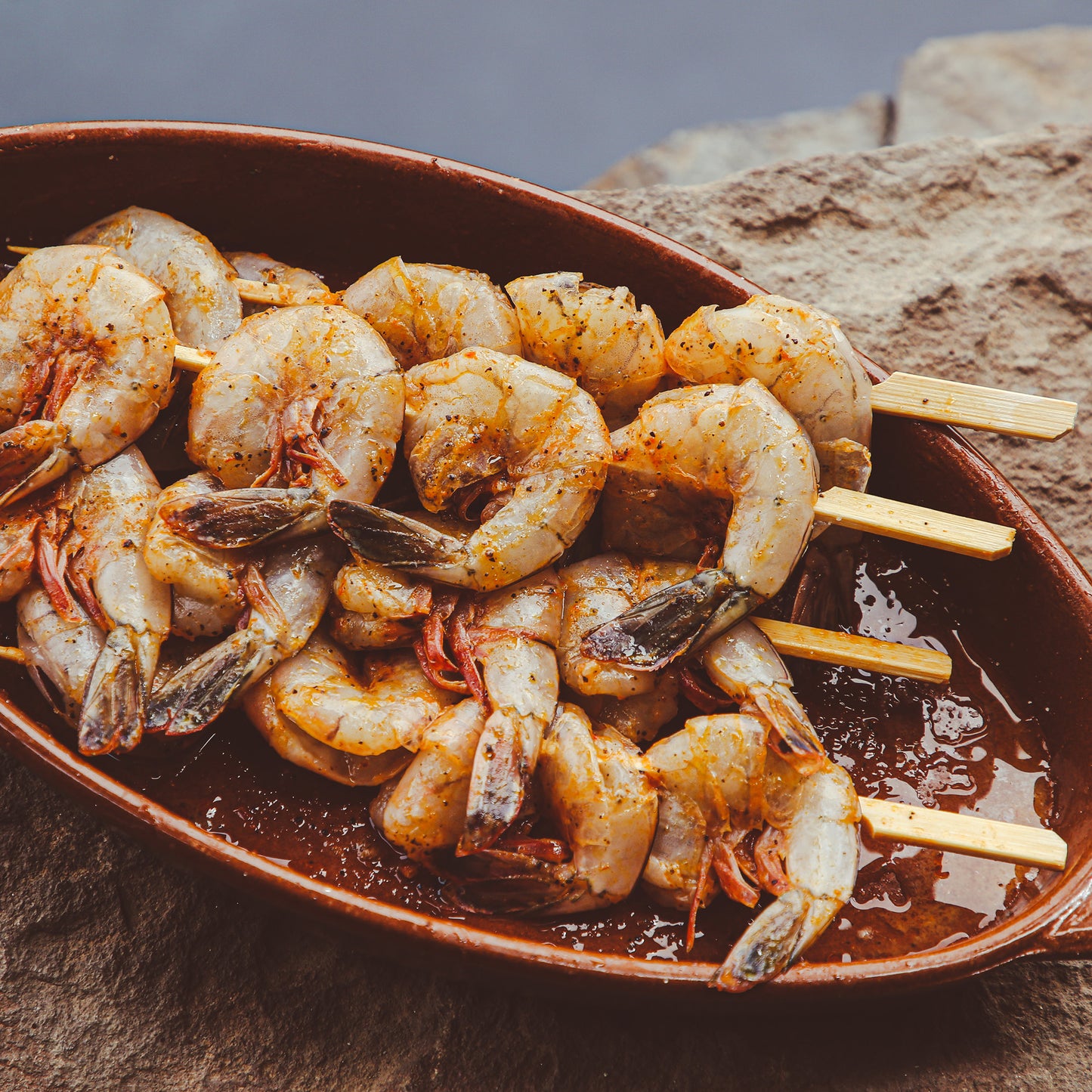 Shrimp on skewers marinated with chile sauce in serving dish on rock ledge.