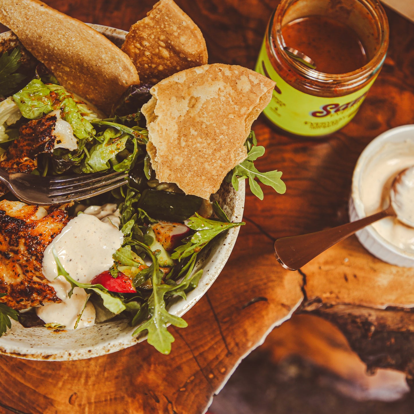 Close-up of fish salad dressed with chile sauce in bowl beside jars on wooden shelf.