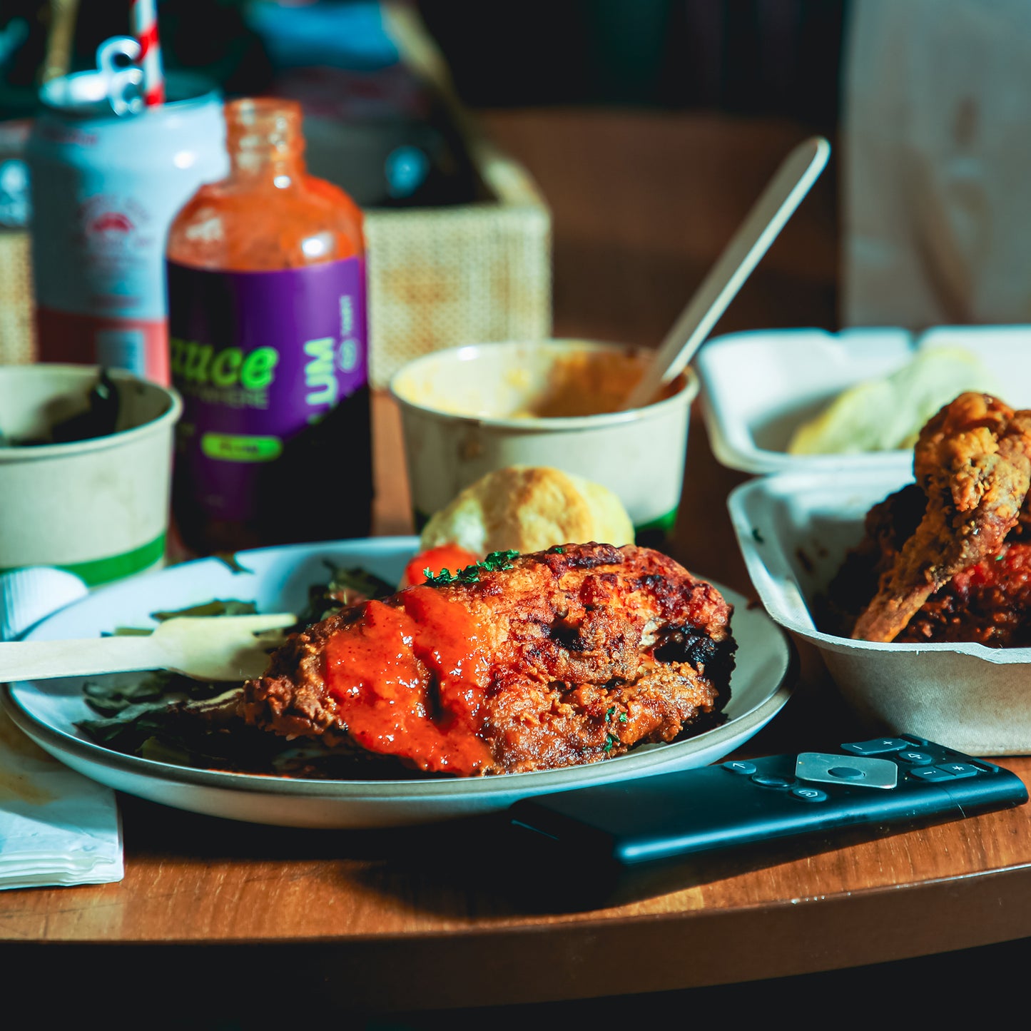 Plate of fried chicken covered in chile sauce beside takeout containers on wooden table.